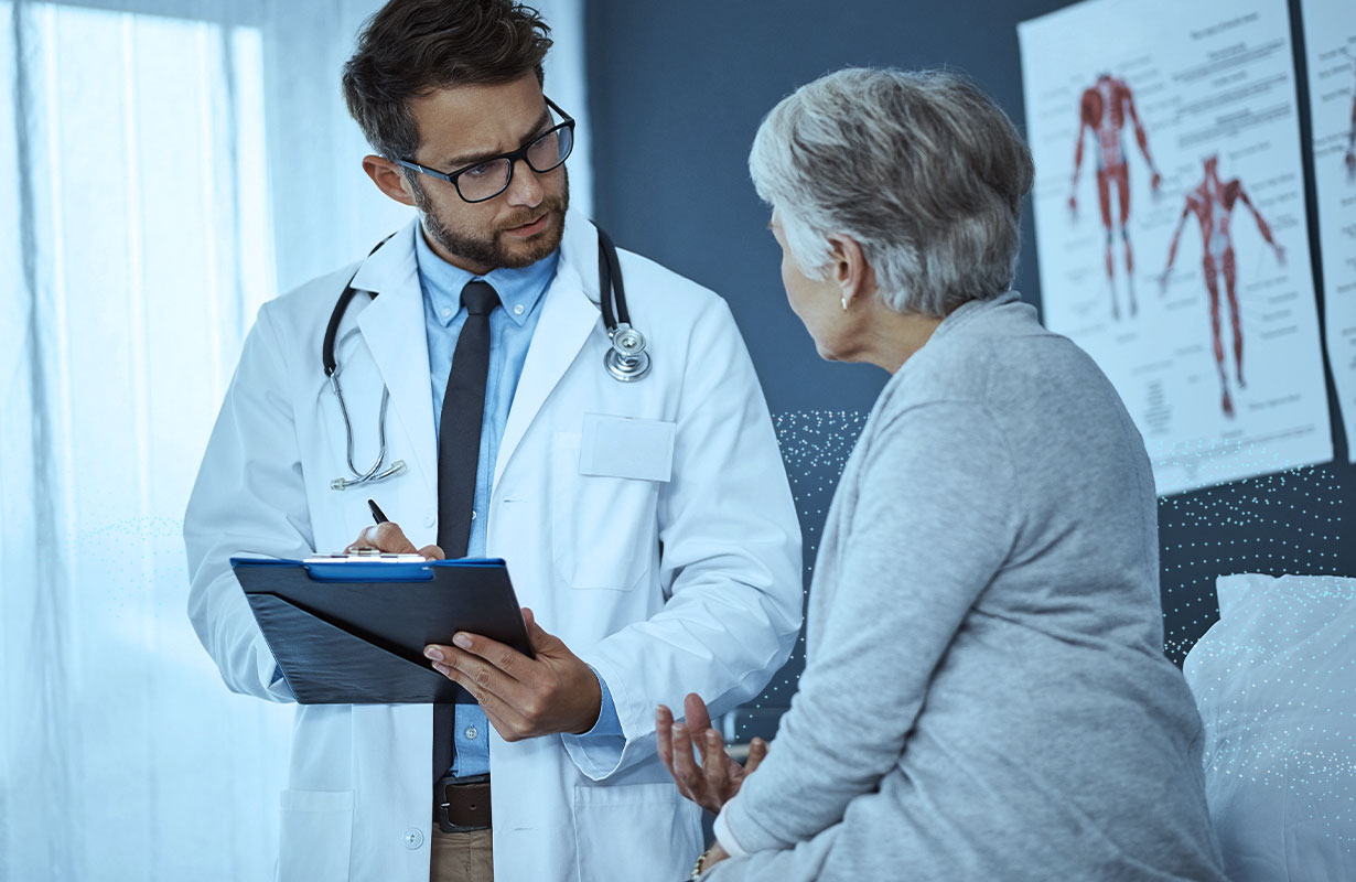 Healthcare professional holding a clipboard whilst talking to an elderly patient in a hospital setting.