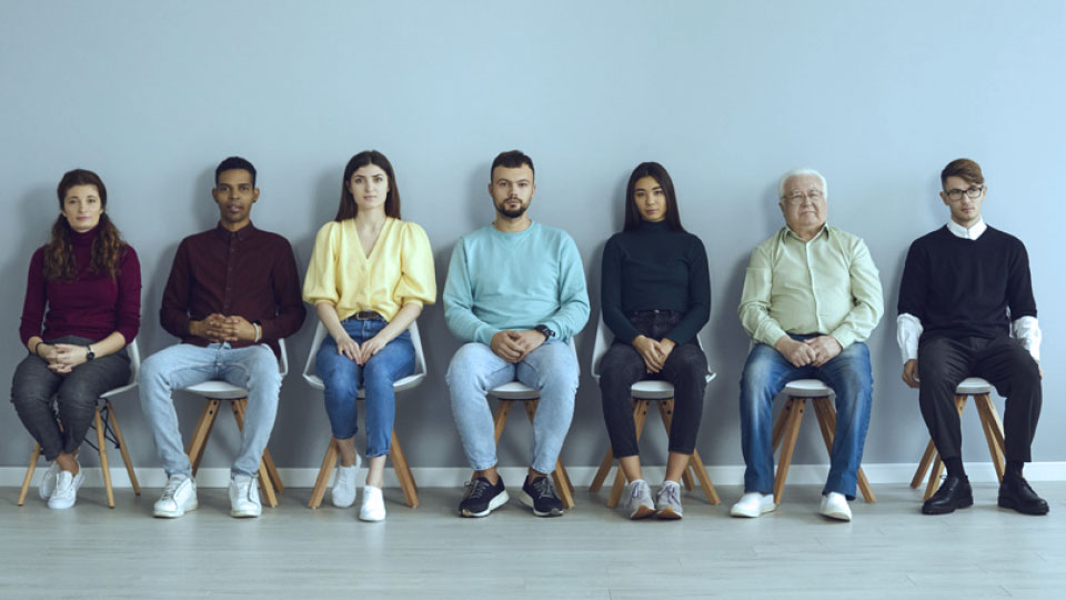 Seven diverse adults sit side by side on stools against a light blue wall.