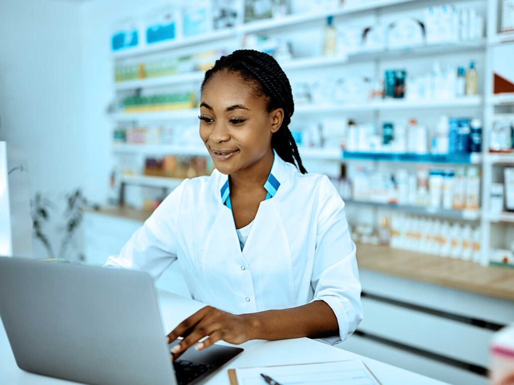 Pharmacist in a white coat using a laptop at a pharmacy counter, with medicine shelves in the background.