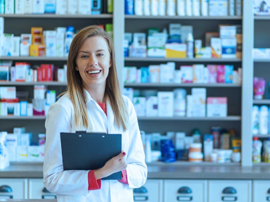 Smiling female pharmacist in a white coat holding a clipboard in front of pharmacy shelves.