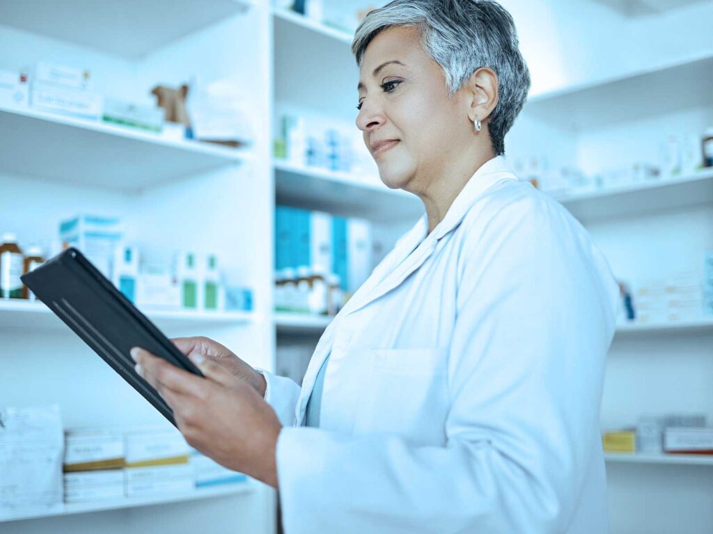 Pharmacist in a white coat reviews a tablet in a pharmacy, with medicine shelves in the background.