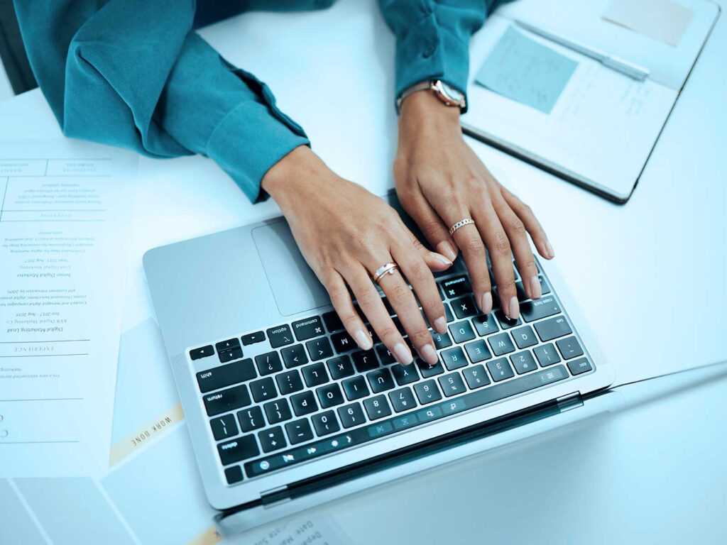Person typing on a laptop at a desk, with documents and a tablet nearby.
