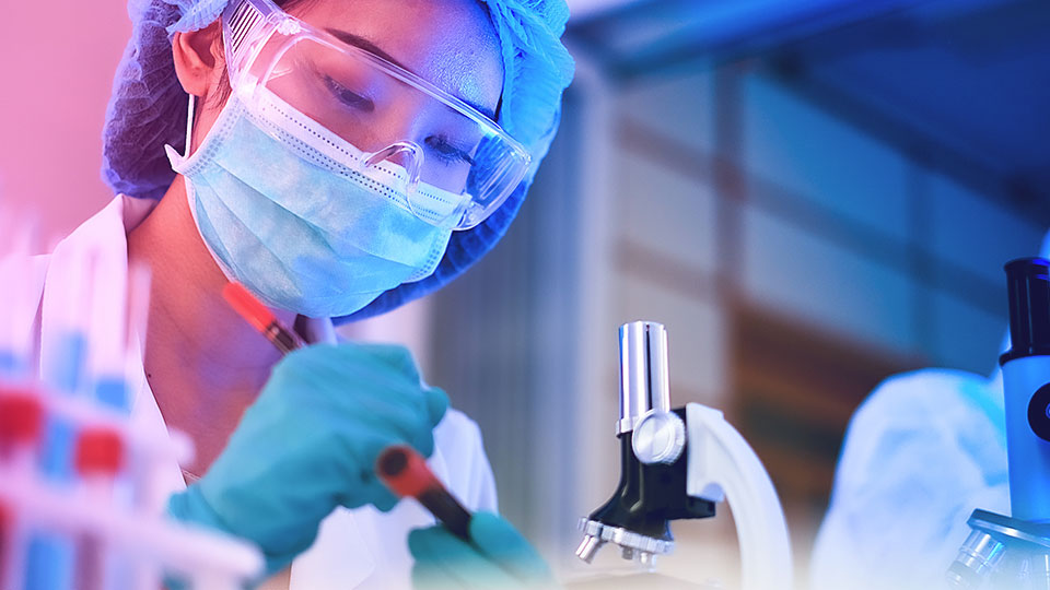 A scientist in a lab coat, mask, and goggles handling a test tube next to a microscope.