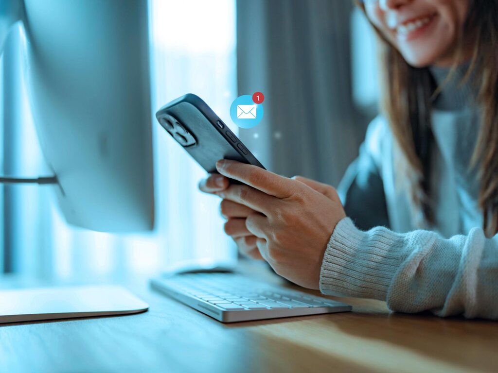 A smiling woman checks her smartphone beside a keyboard at a bright desk, as a glowing email icon signals one new message; a warm, connected moment.