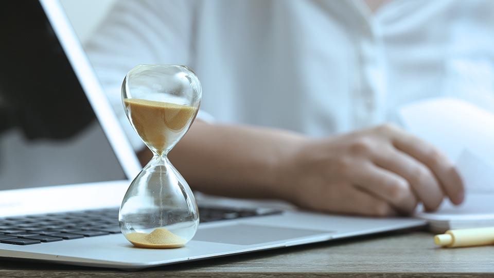 Person working at a desk with a sand timer on a laptop signifying an upcoming deadline.