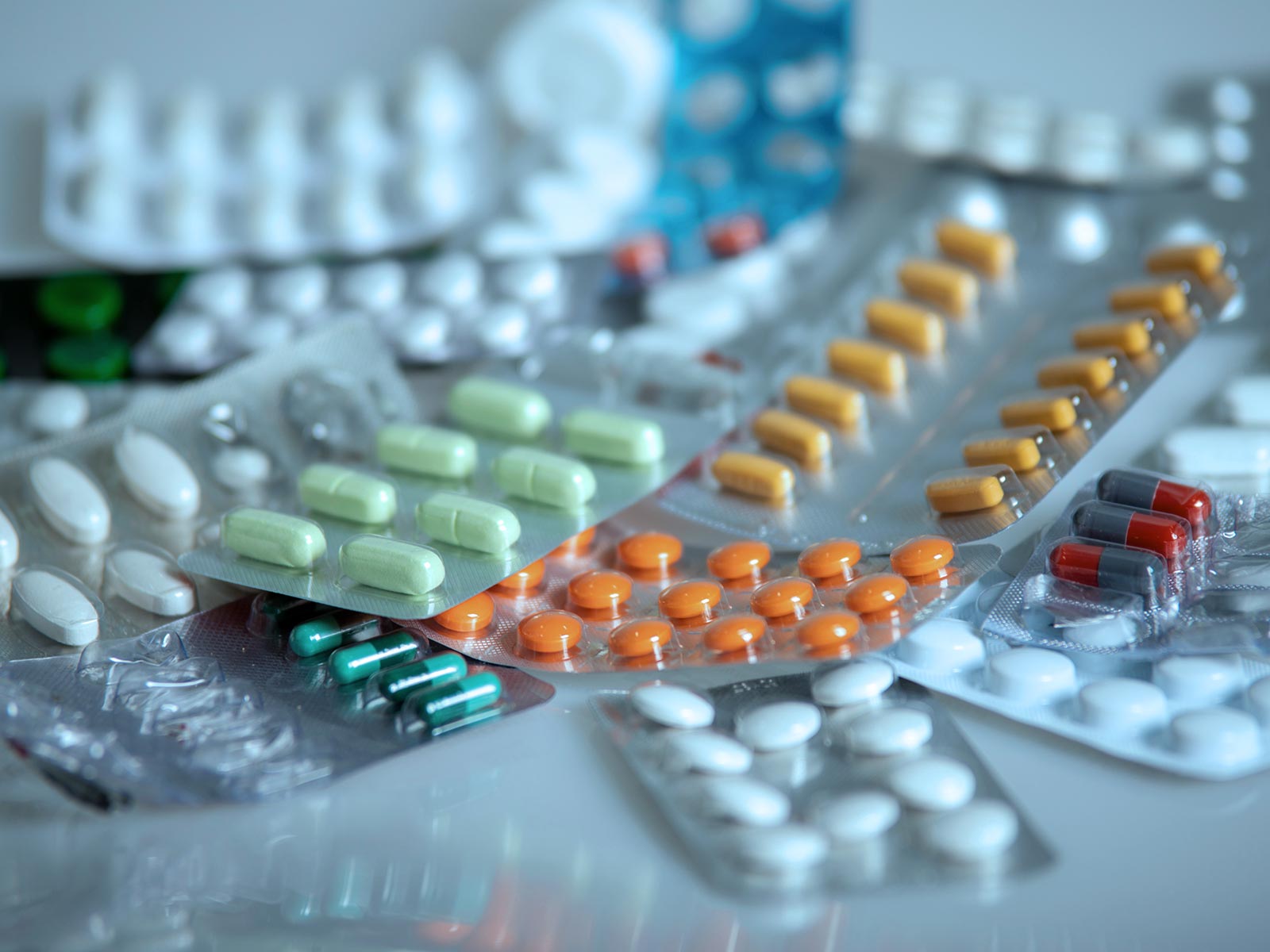 Close-up of colorful blister packs of pills and capsules—orange, yellow, green, red, and white—overlapping on a cool-toned surface, suggesting a clinical pharmacy setting.