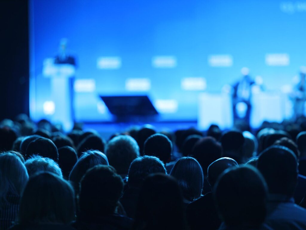 A seated audience watches a speaker on a brightly lit blue conference stage, with a lectern and screen blurred in the background; the mood is attentive and formal.