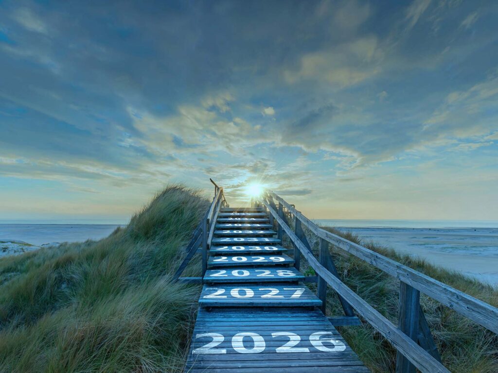 A weathered wooden boardwalk crosses coastal dunes toward the sea, with years painted on its steps and sunlight breaking through dramatic clouds, evoking hope and renewal.