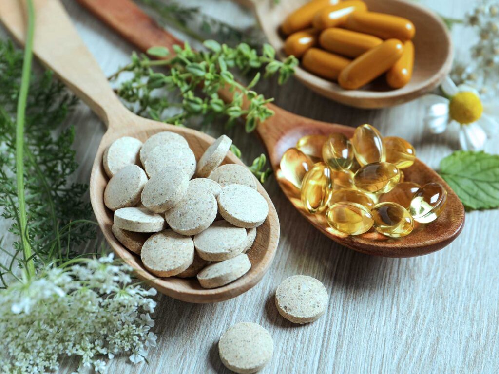 Wooden spoons display pale tablets, amber capsules, and golden oil softgels among fresh herbs and flowers, creating a calm, natural wellness scene.