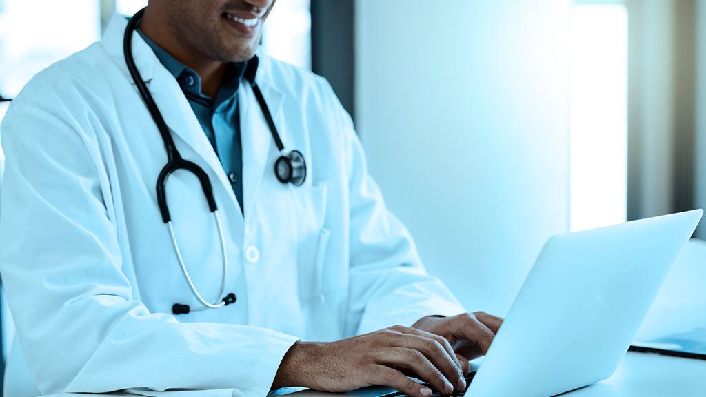 Doctor in a white coat and stethoscope uses a laptop in a bright clinical setting.