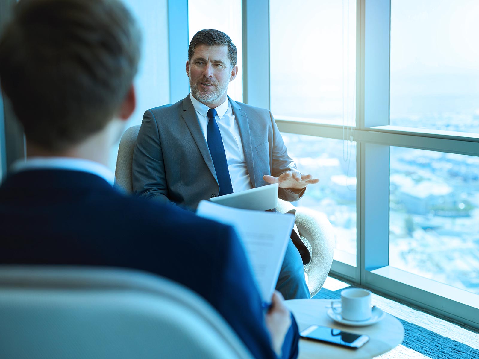 A suited businessman gestures during a meeting with a colleague by large office windows, conveying a calm, professional discussion in a bright setting.