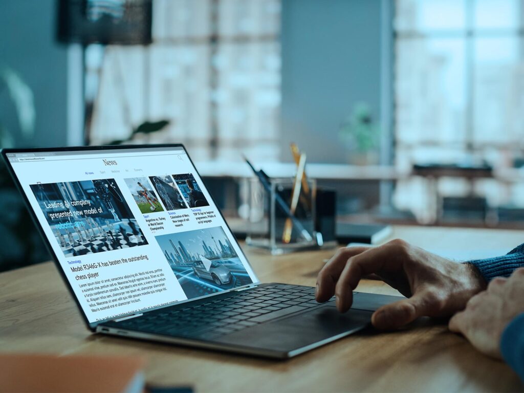 A hand types on a laptop displaying a news website in a bright, modern office, with desk items and large windows creating a calm, focused mood.