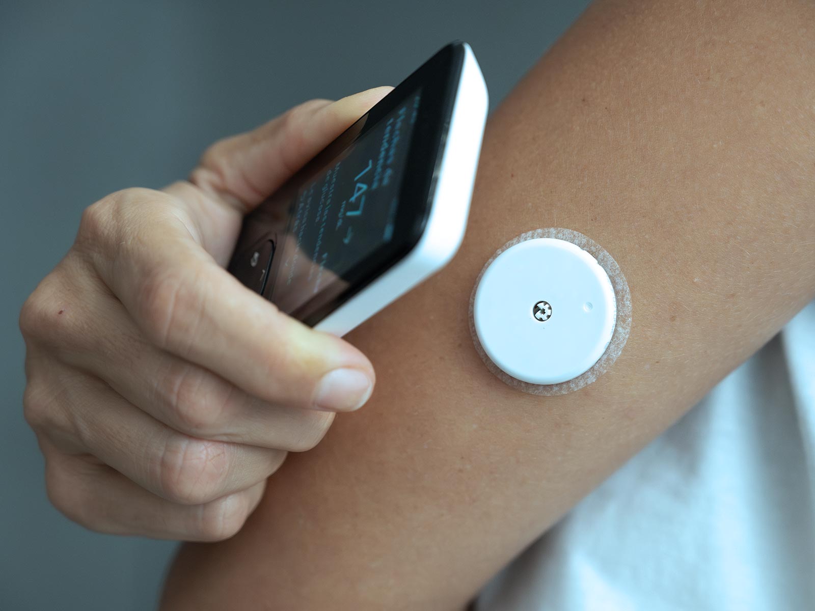 A person holds a smartphone near a round white sensor patch attached to their upper arm, checking health data in a calm, clinical setting.