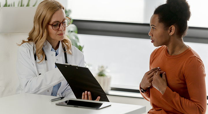 A female doctor in a white coat with a stethoscope sitting at a desk, while a female patient in an orange top sits across from her, holding her hands near her chest.