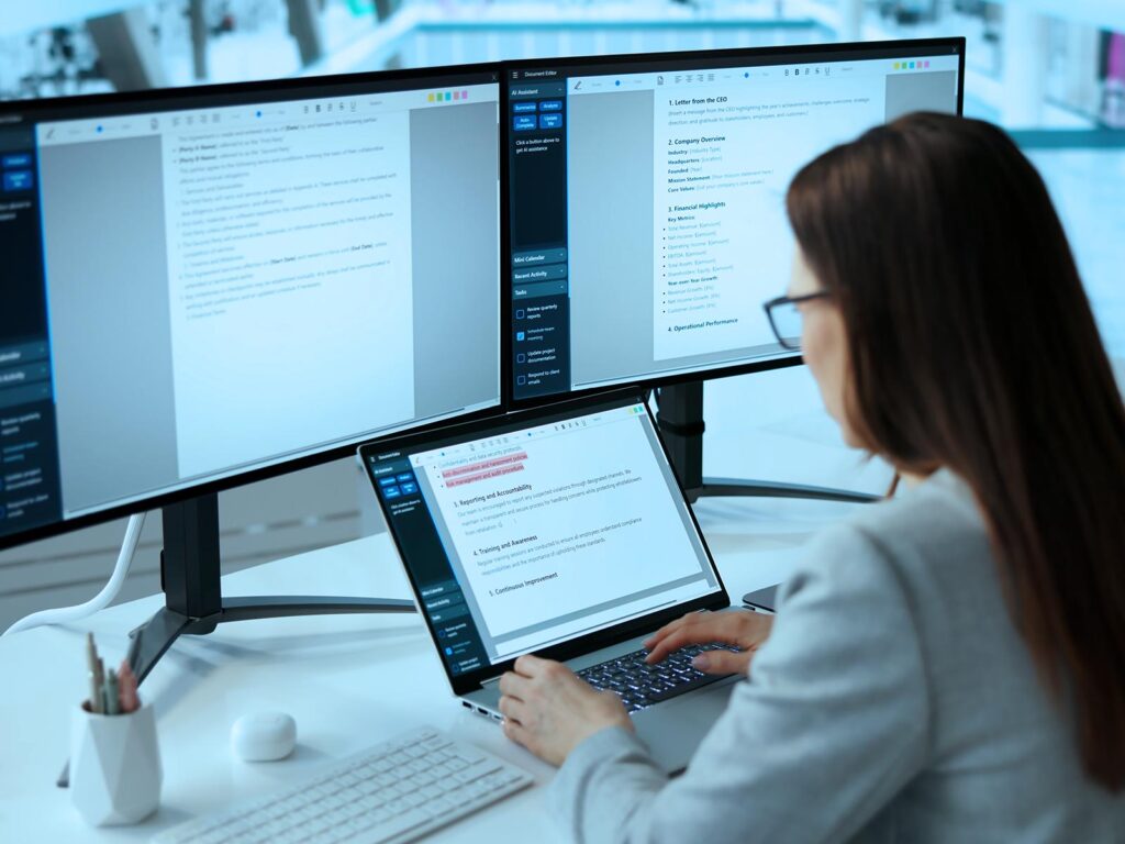 A woman reviews text on two large monitors and a laptop in a bright office, focused on a multi-screen digital document workflow.