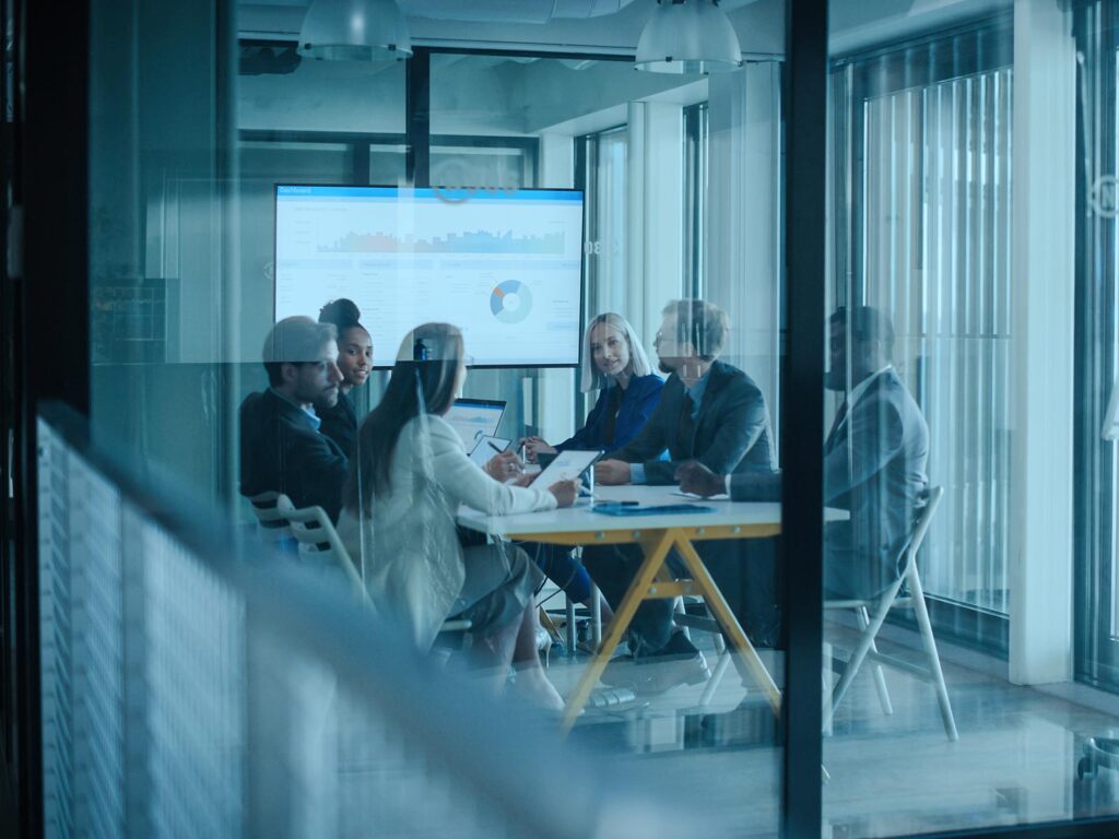 Colleagues meet around a conference table in a glass-walled office, reviewing charts on a screen; cool blue tones create a focused, professional mood.
