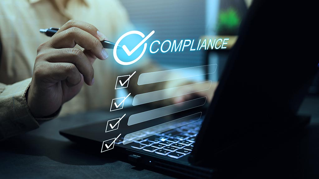 A professional marks a digital compliance checklist beside a laptop, with several boxes checked; the image conveys accuracy, oversight, and workplace accountability.