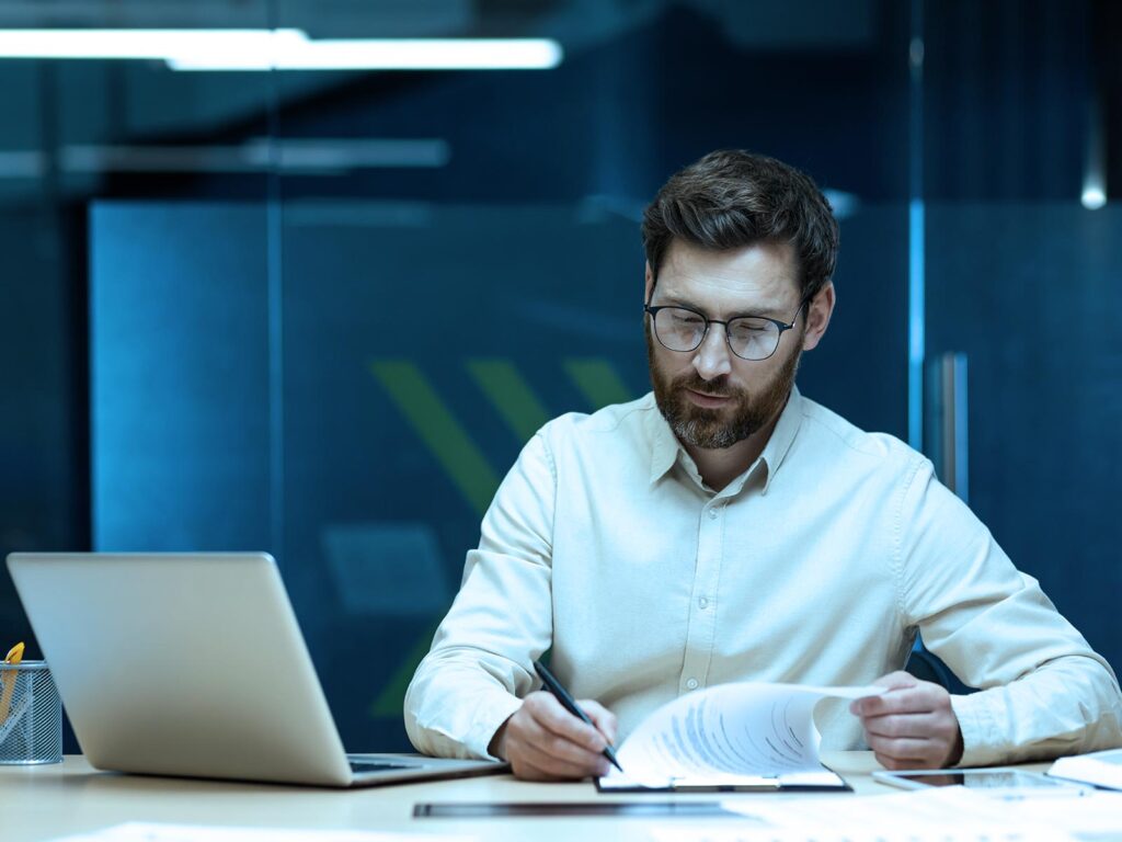 Man with glasses sits at a desk, reviewing and signing documents beside a laptop in an office setting.