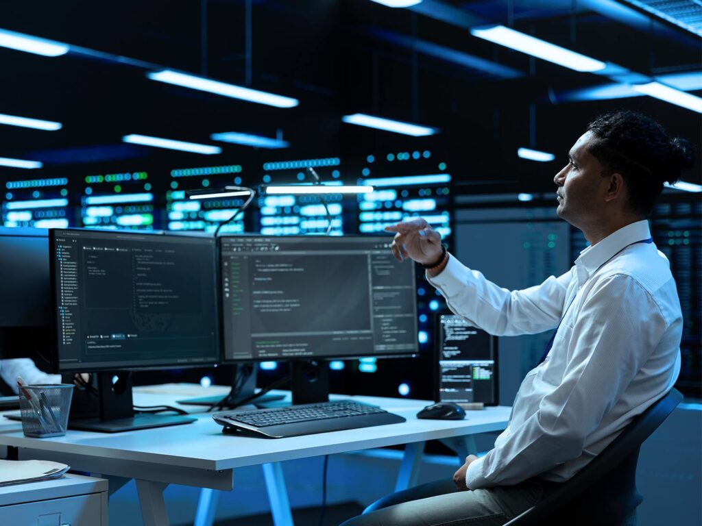 IT professional monitors code and server data on multiple screens in a blue-lit data center.